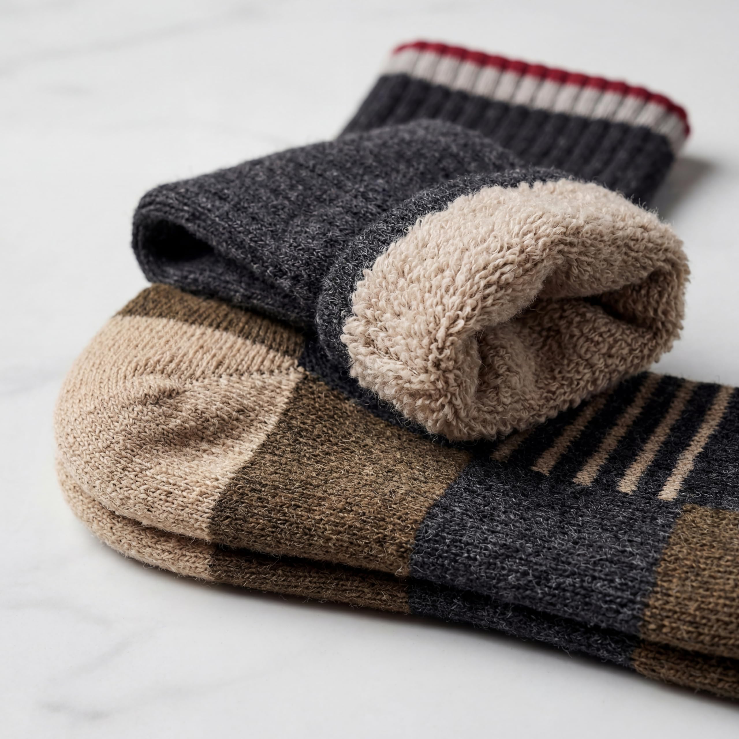 A stack of folded knit socks on a light marble surface. The top sock is dark charcoal gray with a soft beige fleece-like cuff and a red and cream striped ribbed top edge.