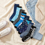 A styled flat-lay of a 9-pack of ankle socks in various patterns and colors, arranged next to light-wash jeans, a cream knit sweater, and white sneakers on a beige fabric background.