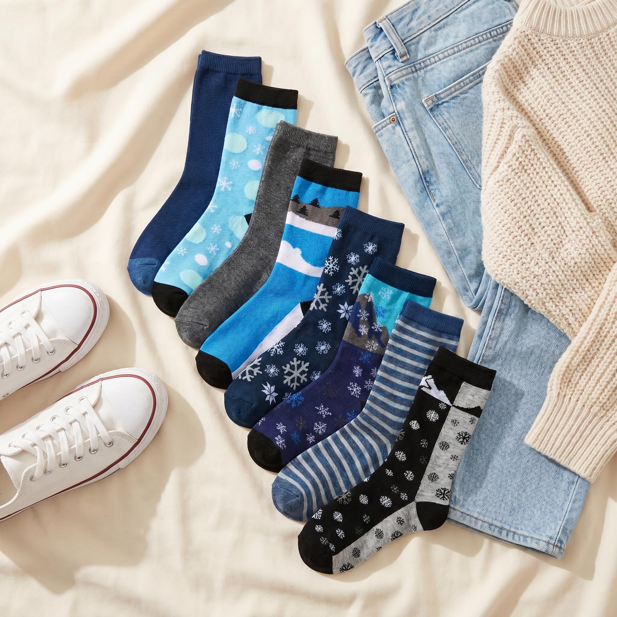 A styled flat-lay of a 9-pack of ankle socks in various patterns and colors, arranged next to light-wash jeans, a cream knit sweater, and white sneakers on a beige fabric background.