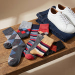 A styled flat-lay of multiple pairs of men's socks in different patterns and colors, arranged on a wooden bench next to folded blue jeans and white sneakers.