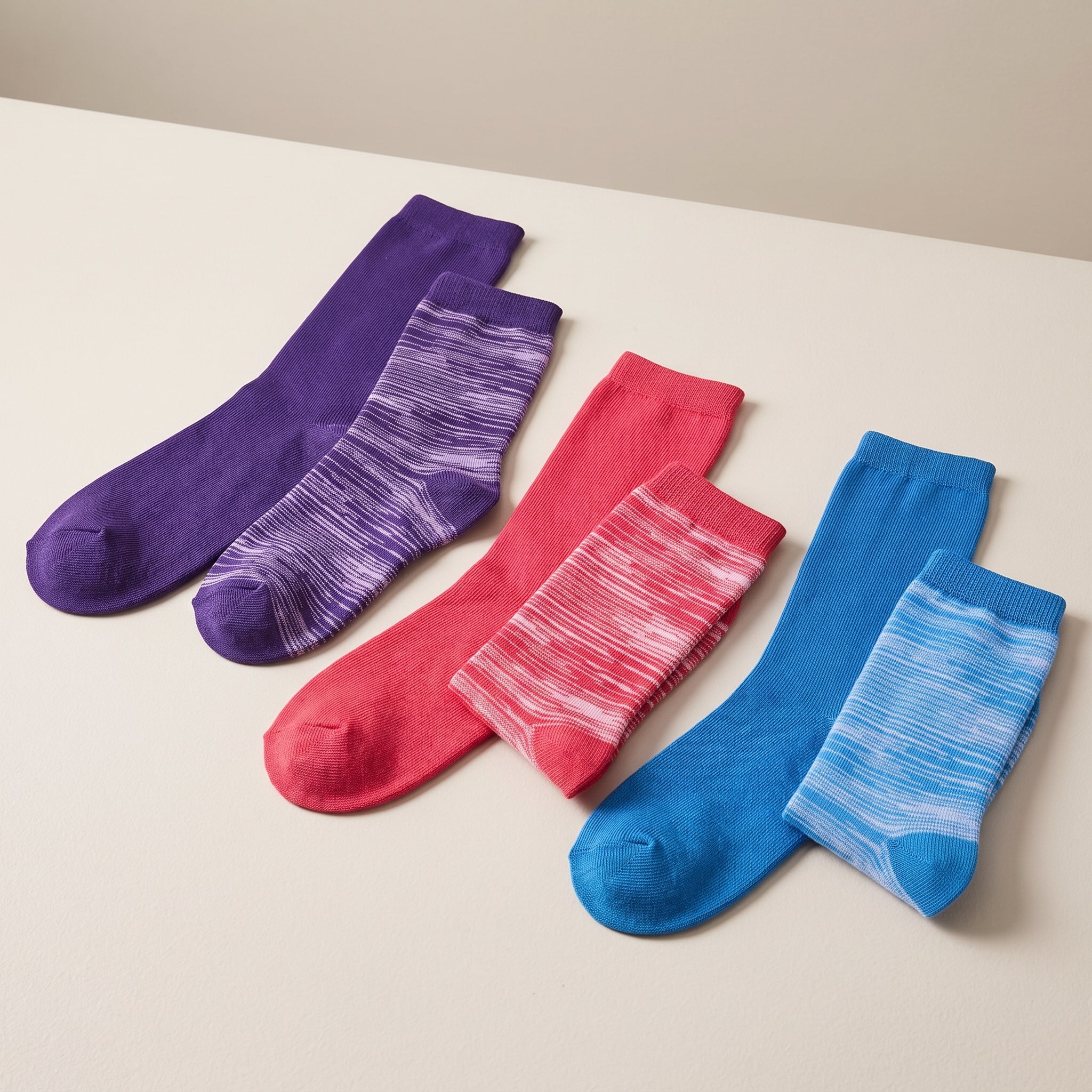 Four pairs of crew socks in purple, pink, and blue, arranged diagonally on a light beige surface. Each pair includes one solid-color sock and one striped sock.