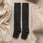 Black knee-high socks on a light surface with a beige scarf, books, and sweater.