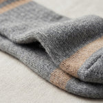 Close-up of a folded knit sock in heather gray with a tan stripe near the cuff, resting on a light beige textured fabric surface.