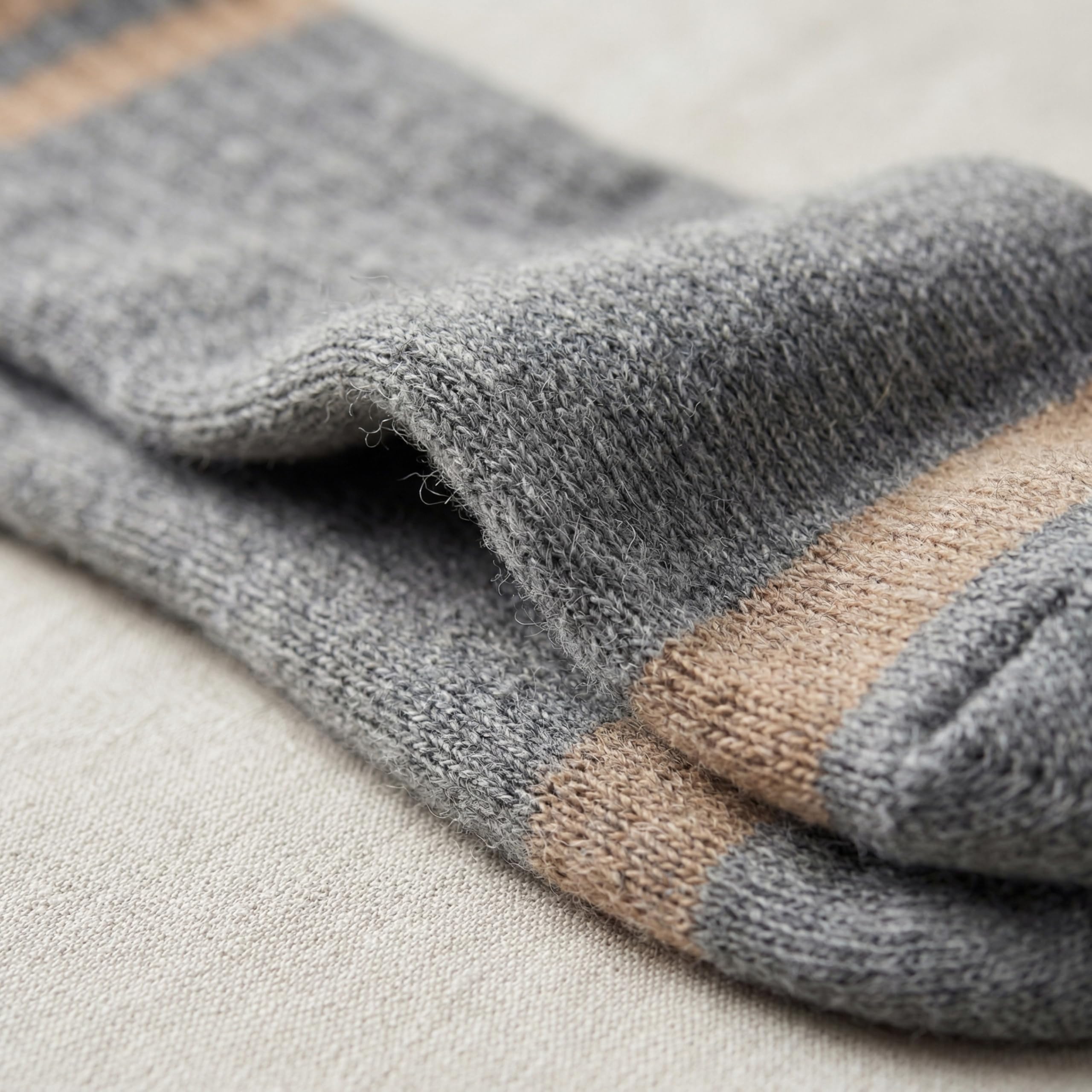 Close-up of a folded knit sock in heather gray with a tan stripe near the cuff, resting on a light beige textured fabric surface.