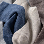 Close-up of folded knit fabric in multiple soft colors, including blue, beige, taupe, and light gray, showing the texture and weave of the material.