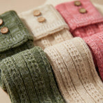 Close-up of folded knit socks or booties in three soft colors: olive green, cream, and dusty pink, each with two wooden buttons near the top.