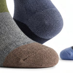 Close-up of three knit socks on feet: one heather gray with a brown toe, one dark blue with a navy heel, and one blue with a green cuff, shown on a white background.