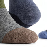 Close-up of three knit socks on feet: one heather gray with a brown toe, one dark blue with a navy heel, and one blue with a green cuff, shown on a white background.