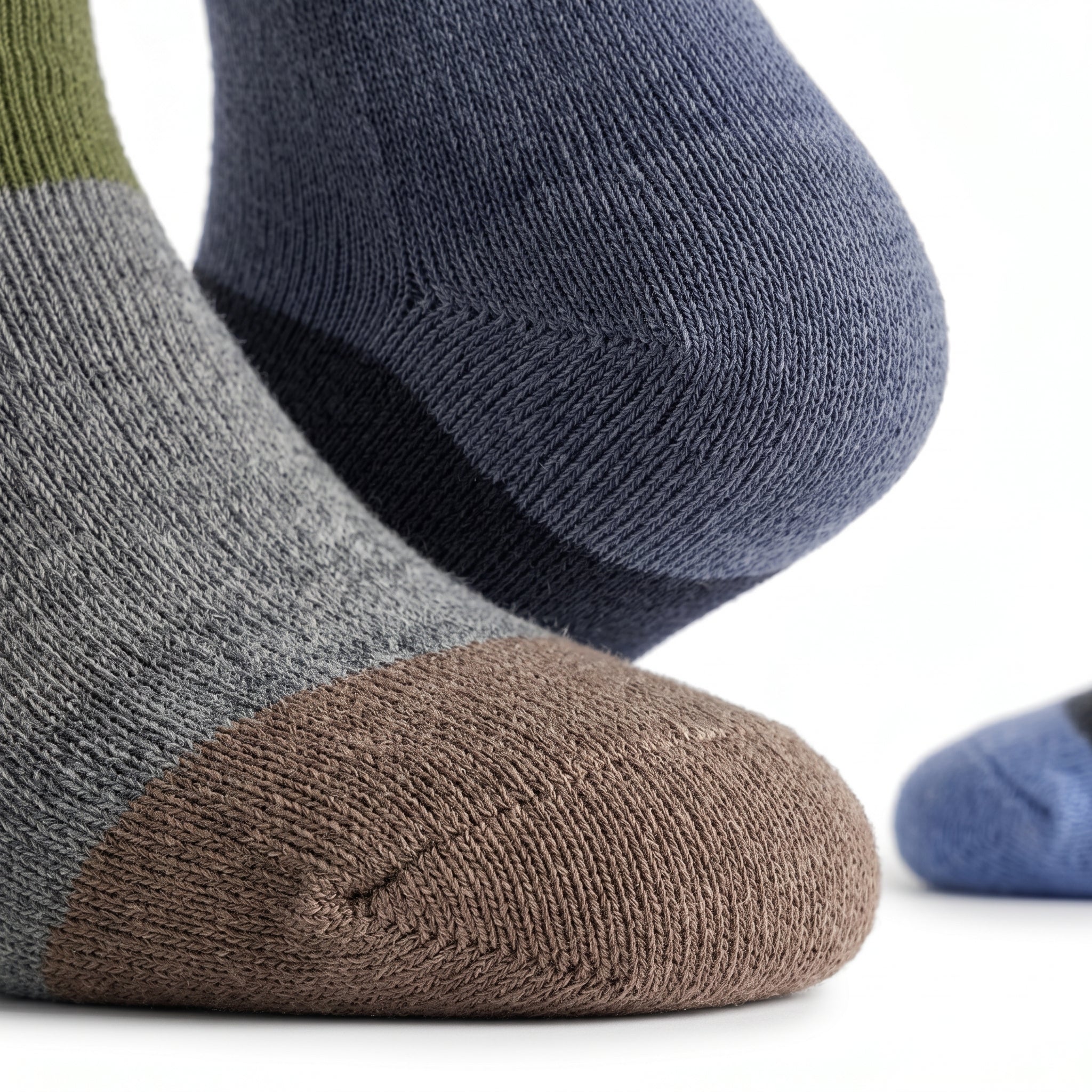 Close-up of three knit socks on feet: one heather gray with a brown toe, one dark blue with a navy heel, and one blue with a green cuff, shown on a white background.