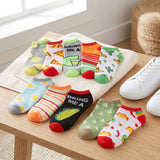 Eight pairs of colorful low-cut ankle socks with various food and pattern designs are arranged on a white towel on a wooden table, next to a white sneaker.