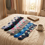 Five pairs of argyle socks in various colors are arranged on a cream cable-knit bedspread in a cozy bedroom setting, accompanied by a vintage book, a ceramic mug, and a clothing rack in the background.