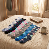 Five pairs of argyle socks in various colors are arranged on a cream cable-knit bedspread in a cozy bedroom setting, accompanied by a vintage book, a ceramic mug, and a clothing rack in the background.