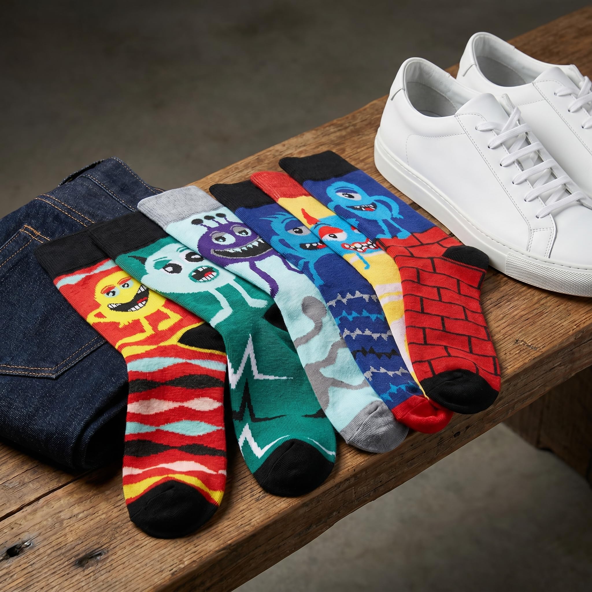 Five pairs of colorful cartoon monster socks arranged on a wooden bench next to folded dark blue jeans and white sneakers. The socks feature various playful monster patterns in bright colors like red, blue, green, yellow, and purple.