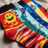 Five pairs of colorful novelty socks with cartoon monster prints, arranged diagonally on a neutral fabric background.