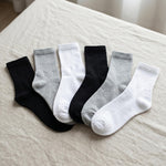 Five pairs of crew socks in black, light gray, white, dark charcoal, and medium gray are arranged in a fan shape on a beige linen surface.