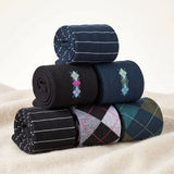 Five rolled pairs of men's dress socks displayed in a stack on a soft beige fabric surface. The socks feature various patterns including navy blue pinstripes, black and navy argyle with pink and blue accents, black and gray argyle with pink lines, green and navy argyle, and navy and green pinstripes.