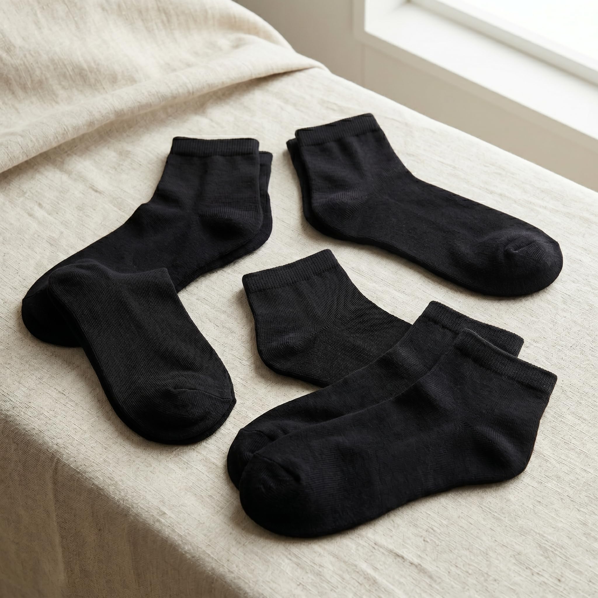 Four pairs of black ankle socks arranged on a light beige linen surface near a window.