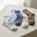 Four pairs of crew socks in light gray, pale blue, medium blue, cream, and taupe brown, arranged on a beige linen surface.