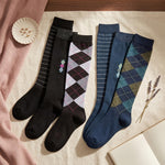 Four pairs of knee-high socks arranged on a cream fabric surface with styled props. The socks include black and navy options with white stripes, plus two argyle patterns in lavender-gray and teal-blue.