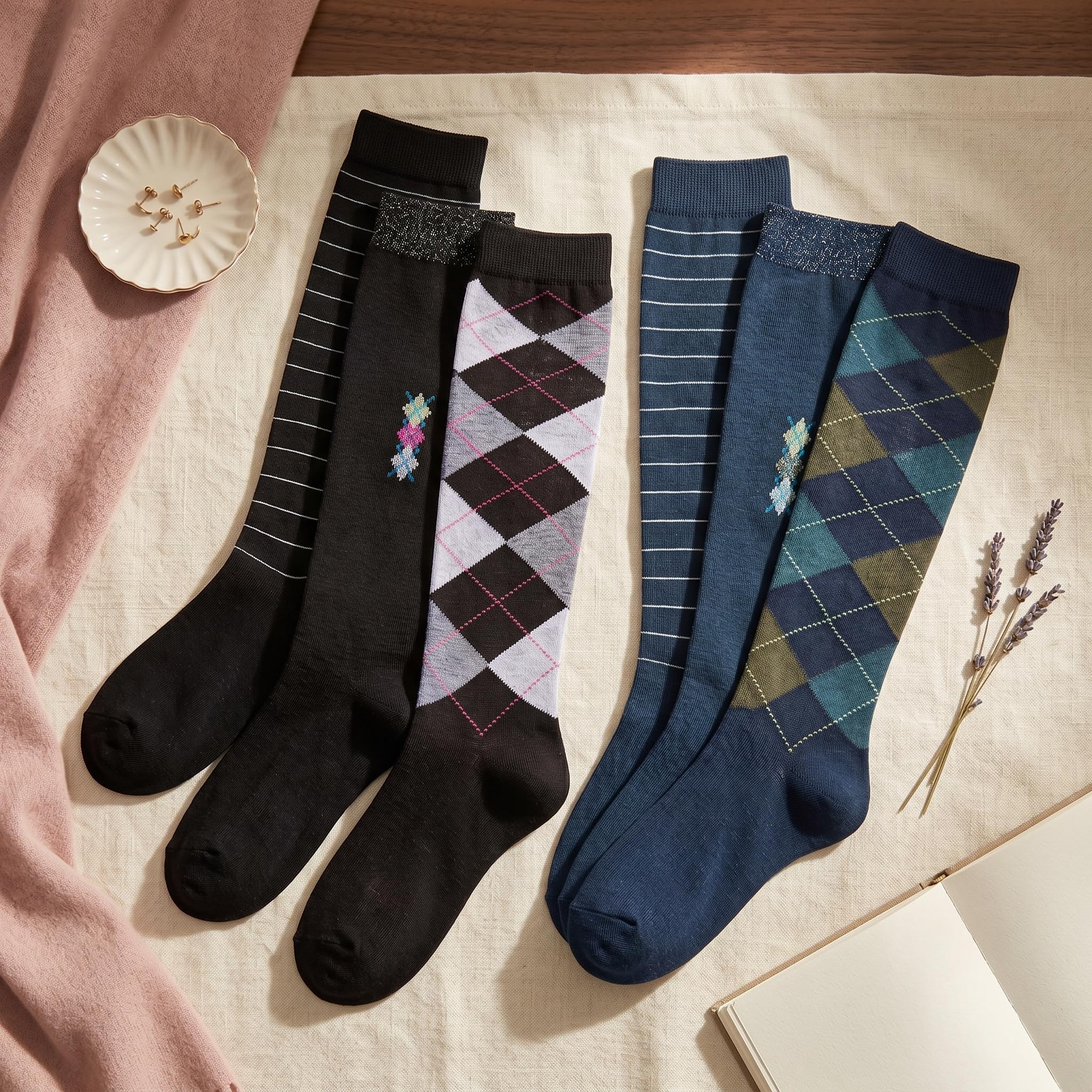 Four pairs of knee-high socks arranged on a cream fabric surface with styled props. The socks include black and navy options with white stripes, plus two argyle patterns in lavender-gray and teal-blue.