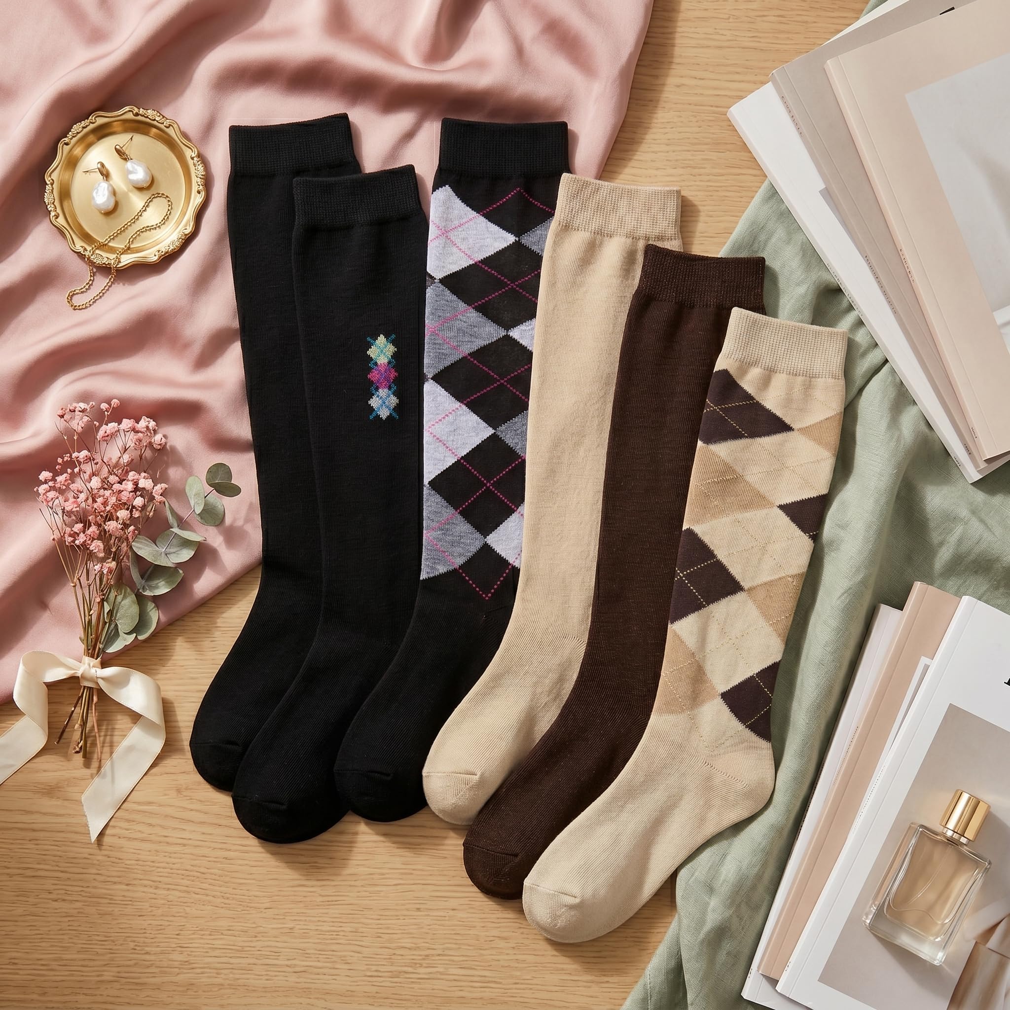 Four pairs of women's knee-high socks arranged in a styled flat lay on a wooden surface with pink fabric, books, dried flowers, and a perfume bottle. The socks include solid black, solid beige, solid brown, and argyle patterns in black, gray, and brown.