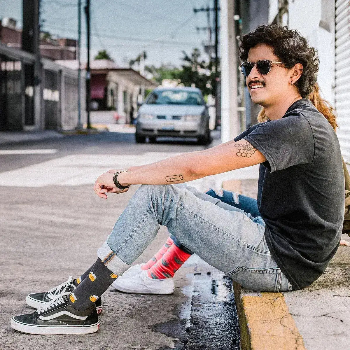 Person sitting on a street curb wearing sunglasses, black shirt, jeans, and colorful socks.
