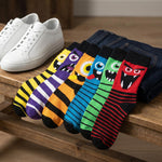 Six pairs of colorful monster-patterned crew socks arranged on a wooden bench next to folded dark blue jeans and a pair of white sneakers.