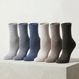 Six pairs of crew socks in different neutral colors are arranged in a row on a light concrete pedestal against a plain off-white background. The colors include light gray, medium gray, slate blue, cream, taupe, and dark charcoal.