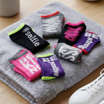 Six pairs of folded ankle socks in assorted colors and patterns, featuring motivational text like '#selfie', 'BE KING', and 'BE positive', arranged on a gray towel with a white sneaker partially visible in the corner.