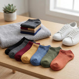 Six pairs of low-cut ankle socks in different colors are arranged on a light wood table next to a stack of folded towels and a pair of white sneakers. The socks include black, gray, burgundy, mustard yellow, blue, olive green, and terracotta orange.