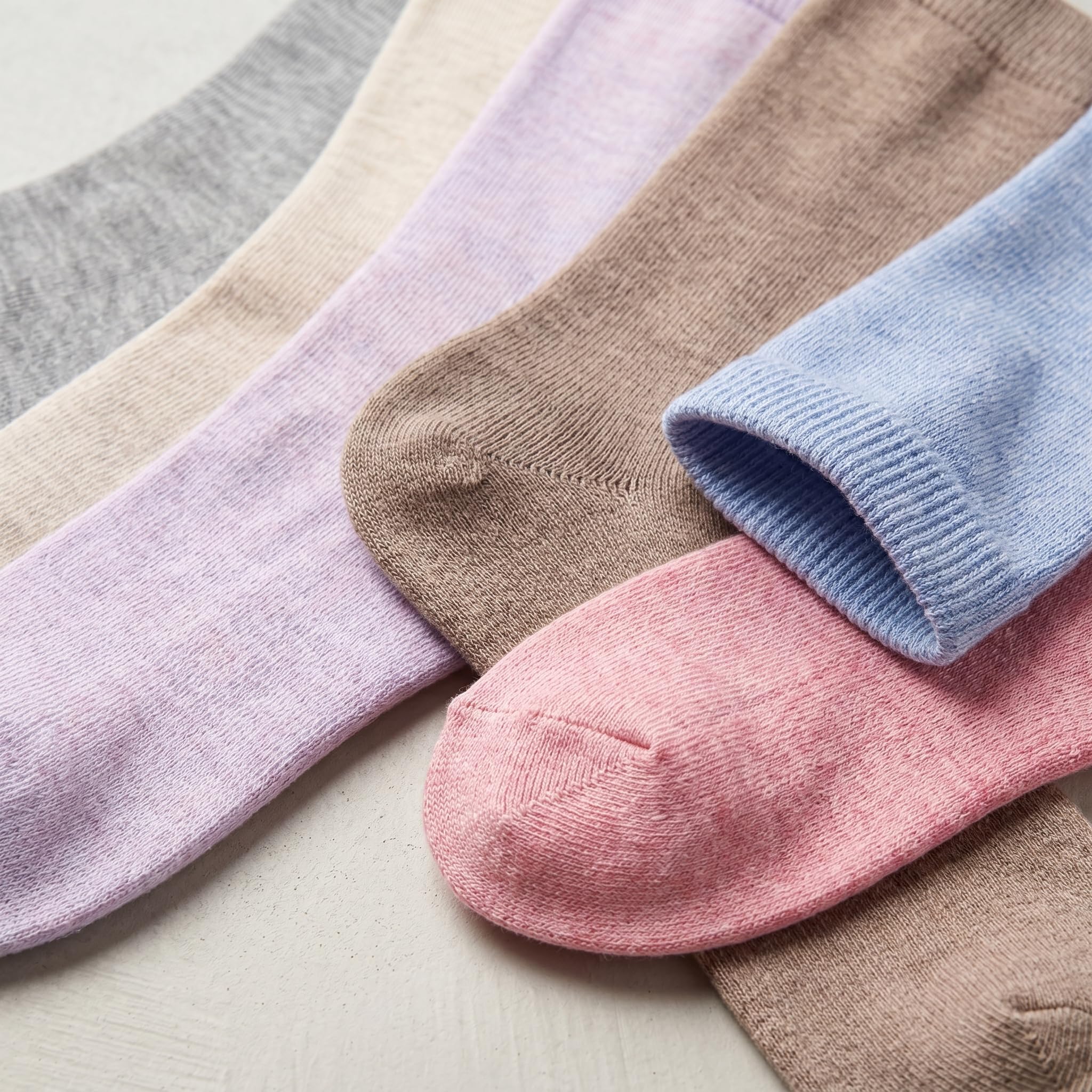 Six pairs of ribbed ankle socks in soft pastel and neutral tones, including gray, cream, lavender, taupe, light blue, and pink, arranged diagonally on a light textured surface.