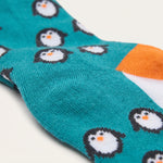 A close-up of a teal knit sock with a repeating penguin pattern, featuring white, black, and orange penguins, and an orange and white toe section.