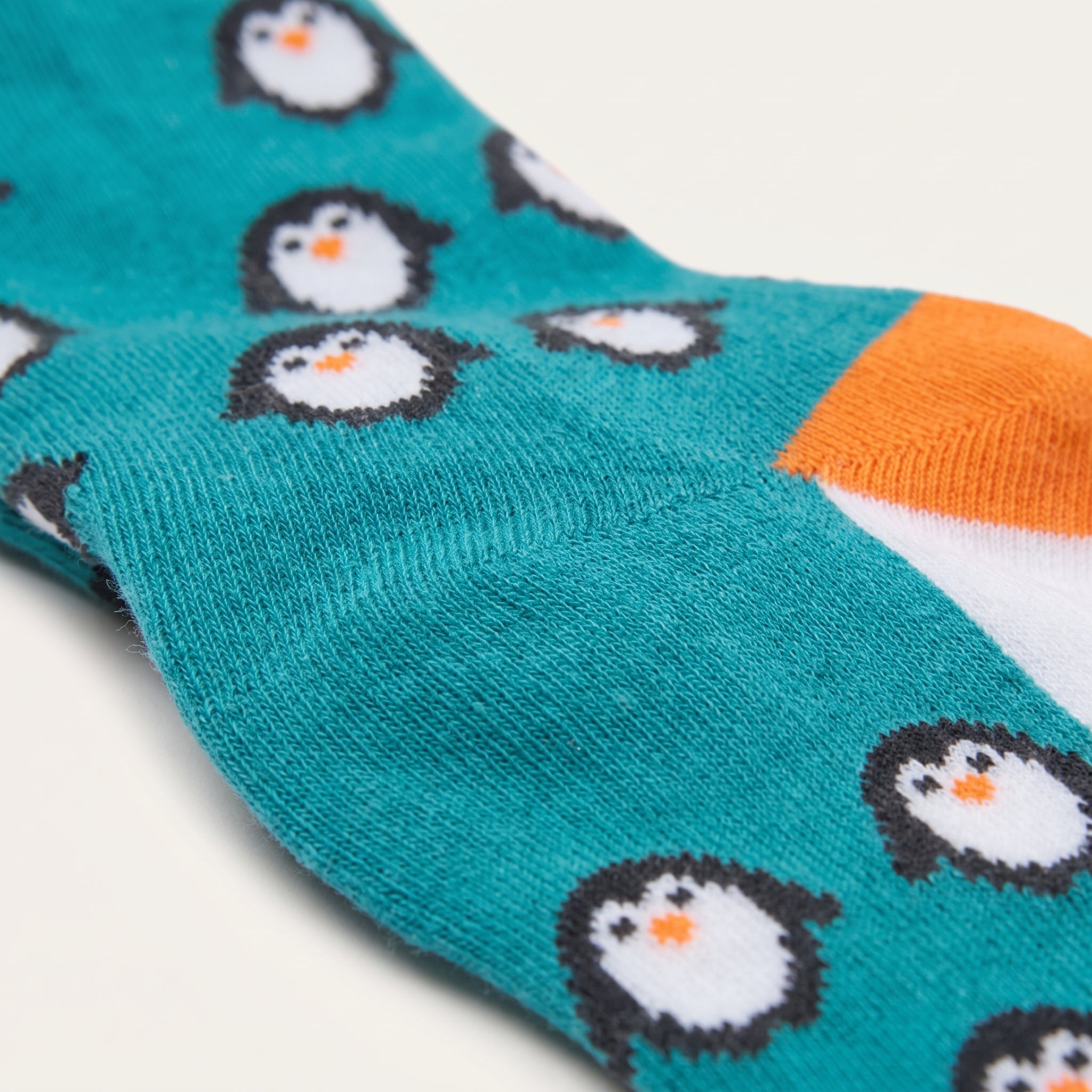 A close-up of a teal knit sock with a repeating penguin pattern, featuring white, black, and orange penguins, and an orange and white toe section.