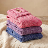Three folded knit socks stacked on a cream cable-knit blanket, with a pink pair on top featuring two gold buttons, followed by purple and blue pairs.