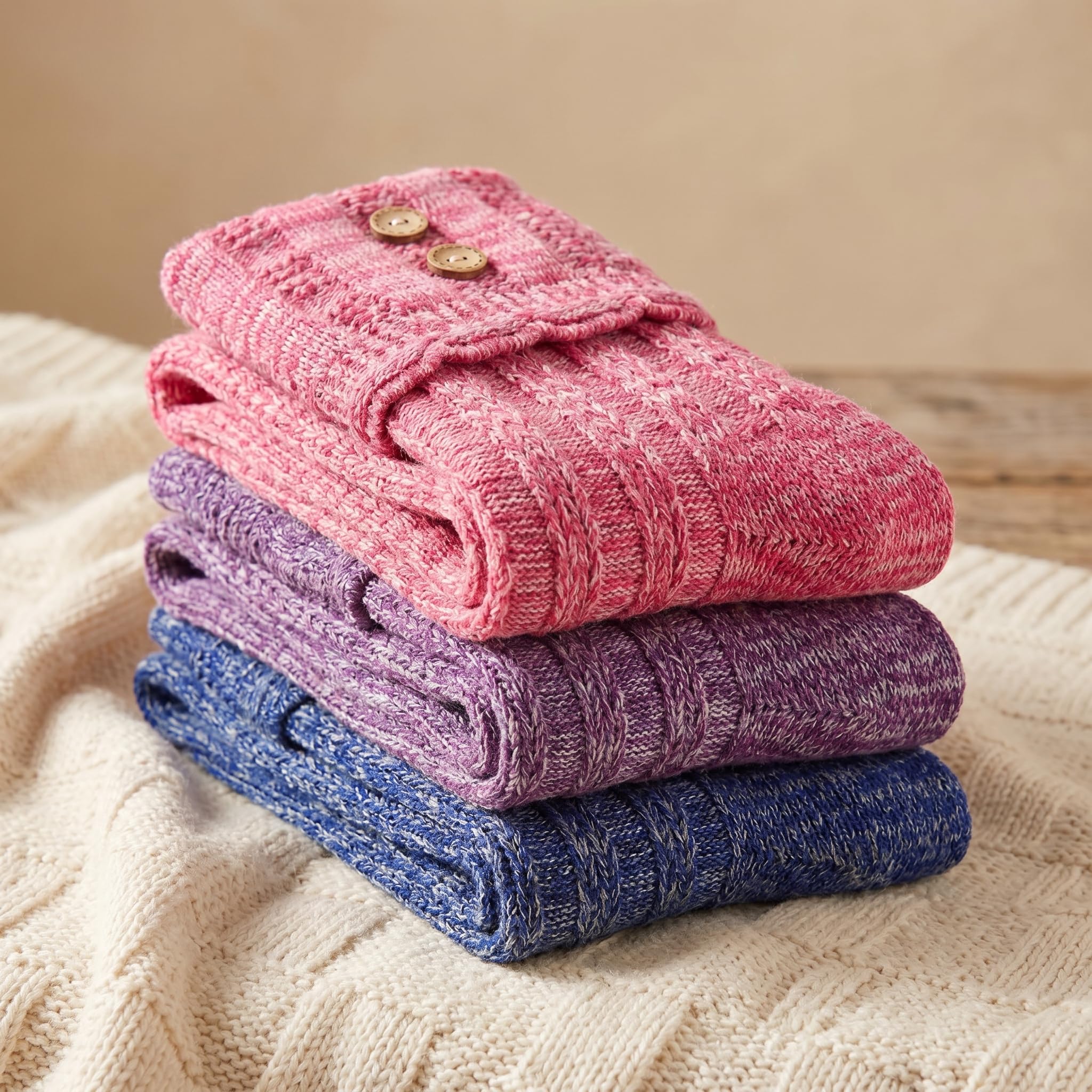 Three folded knit socks stacked on a cream cable-knit blanket, with a pink pair on top featuring two gold buttons, followed by purple and blue pairs.