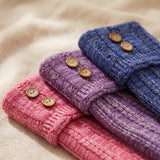 Three folded ribbed knit fabric cuffs in pink, purple, and blue, each decorated with two round wooden buttons, arranged diagonally on a soft beige fabric background.