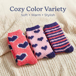 Three pairs of cozy fuzzy socks displayed on a cream-colored knit blanket, with text overlay reading 'Cozy Color Variety' and 'Soft • Warm • Stylish'. The socks feature different patterns: pink with dark purple hearts, light pink with dark purple hearts, and dark purple with coral-pink and white stripes.