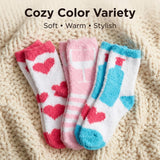 Three pairs of cozy fuzzy socks displayed on a cream knit blanket, with text at the top reading 'Cozy Color Variety' and 'Soft • Warm • Stylish'. The socks feature playful patterns: red hearts on white, pink stripes and wine glass on pink, and blue bottle and cloud shapes on white.