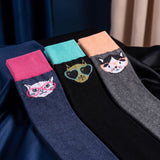 Three pairs of novelty socks with cartoon cat faces are arranged on a dark satin fabric. The socks are navy blue with a pink cuff and a white cat face wearing pink glasses; black with a turquoise cuff and a gold cat face wearing black sunglasses; and dark gray with a peach cuff and an orange-and-black cat face.