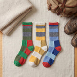 Three pairs of striped crew socks laid flat on a light fabric surface, styled with a folded beige textile, a tan leather bag, and brown boots nearby. The socks feature multicolor horizontal and block stripe patterns in green, red, yellow, gray, blue, and white.