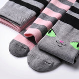 Three pairs of striped socks arranged on a white background. One pair is gray and pink with black stripes, another is pink and gray with black stripes, and the front pair is gray with a cute embroidered cat face in green, black, and pink.