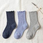 Three ribbed ankle socks with ruffled edges in navy blue, light periwinkle blue, and heather gray, arranged on a textured off-white fabric background with a small decorative paper clip and dried flower.