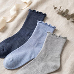 Three ribbed ankle socks with scalloped edges in navy, light blue, and heather gray, arranged diagonally on a beige fabric background with ribbon and eucalyptus props.