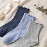 Three ribbed ankle socks with scalloped edges in navy, light blue, and heather gray, arranged diagonally on a beige fabric background with ribbon and eucalyptus props.