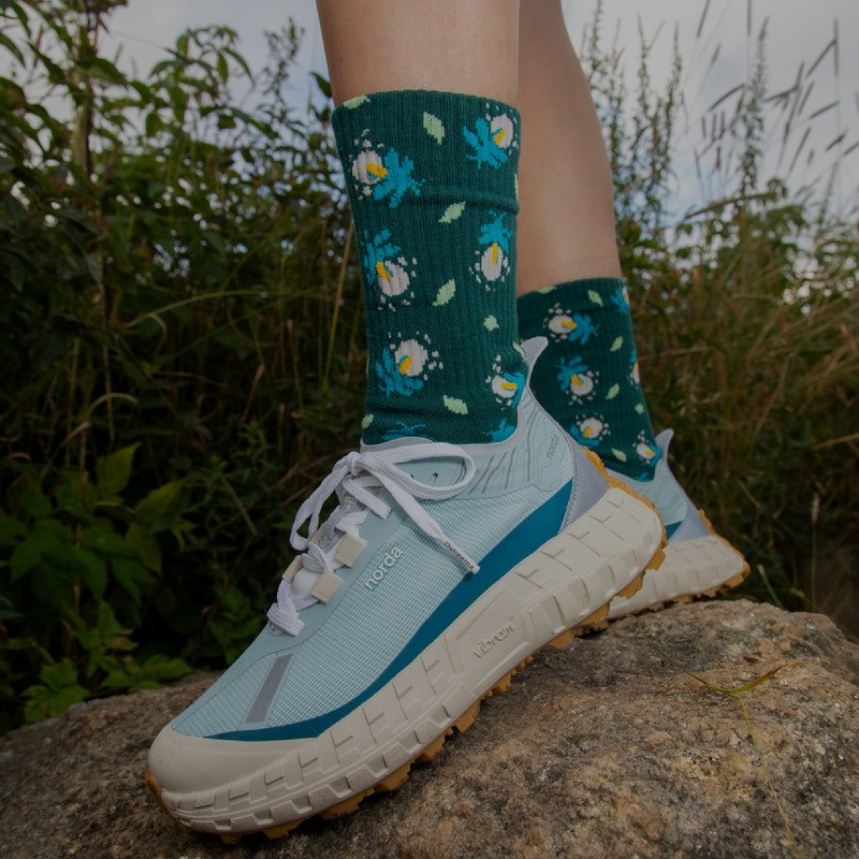 Person wearing blue and white sneakers with green socks featuring floral patterns, standing on a rock with a natural background.