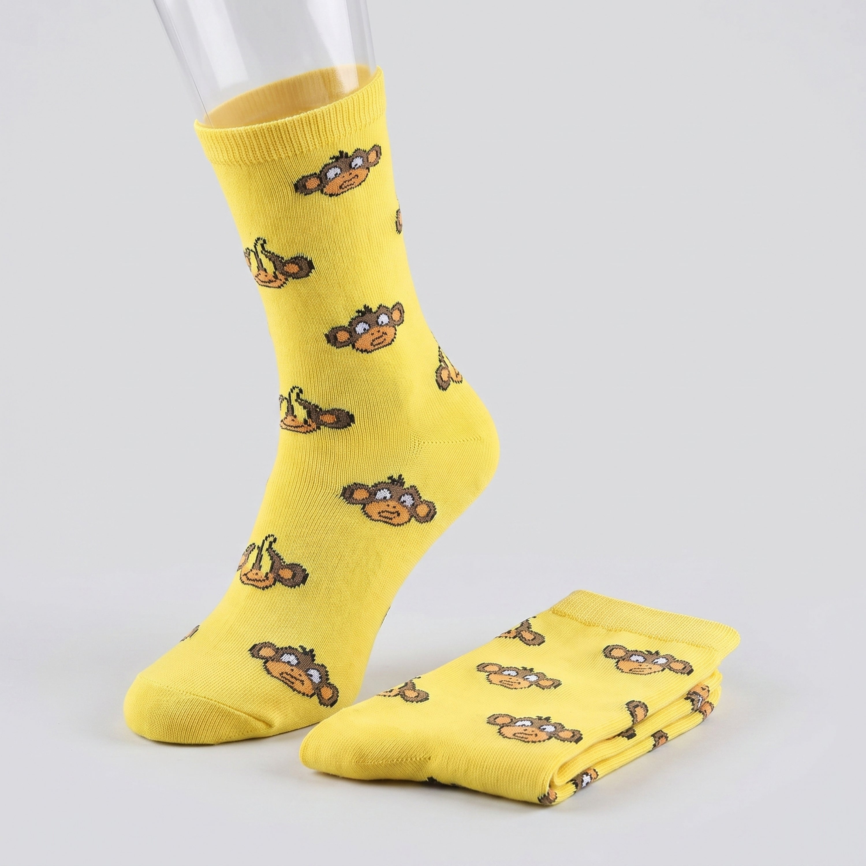Yellow crew socks with a repeating cartoon monkey face pattern, shown on a mannequin foot with one folded pair on a light gray background.