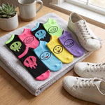 Six pairs of ankle socks in black, pink, blue, green, yellow, and purple, each printed with a different cartoon face, arranged on a gray towel alongside white sneakers on a wooden table.