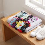 A set of colorful cartoon-patterned baby socks arranged on folded gray towels on a wooden bench, with a pair of white sneakers beside them in a bright indoor setting.