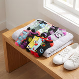 A set of colorful cartoon-patterned baby socks arranged on folded gray towels on a wooden bench, with a pair of white sneakers beside them in a bright indoor setting.
