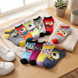 A set of colorful children's ankle socks with cartoon monster faces, arranged on a beige towel on a wooden table next to white sneakers and a small potted succulent.